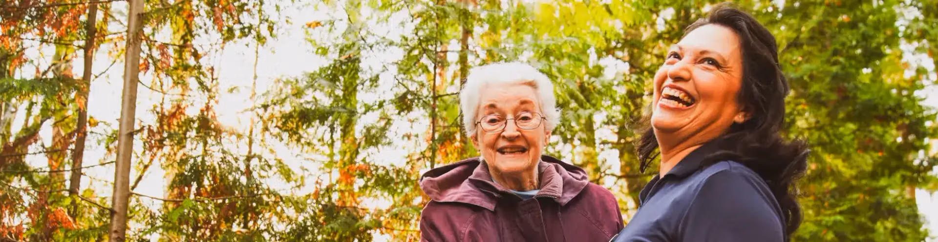 A resident and companion laughing together on an autumn woodland walk near an Áegis Living community