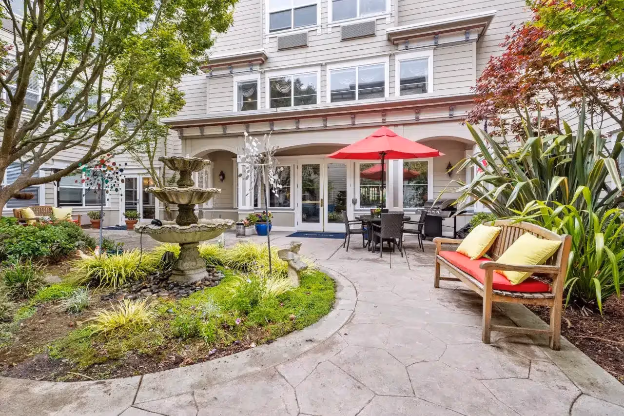 An older woman and a warm caregiver walking together through a sunlit garden courtyard at a memory care in Burlingame, CA community on a mild Peninsula afternoon.