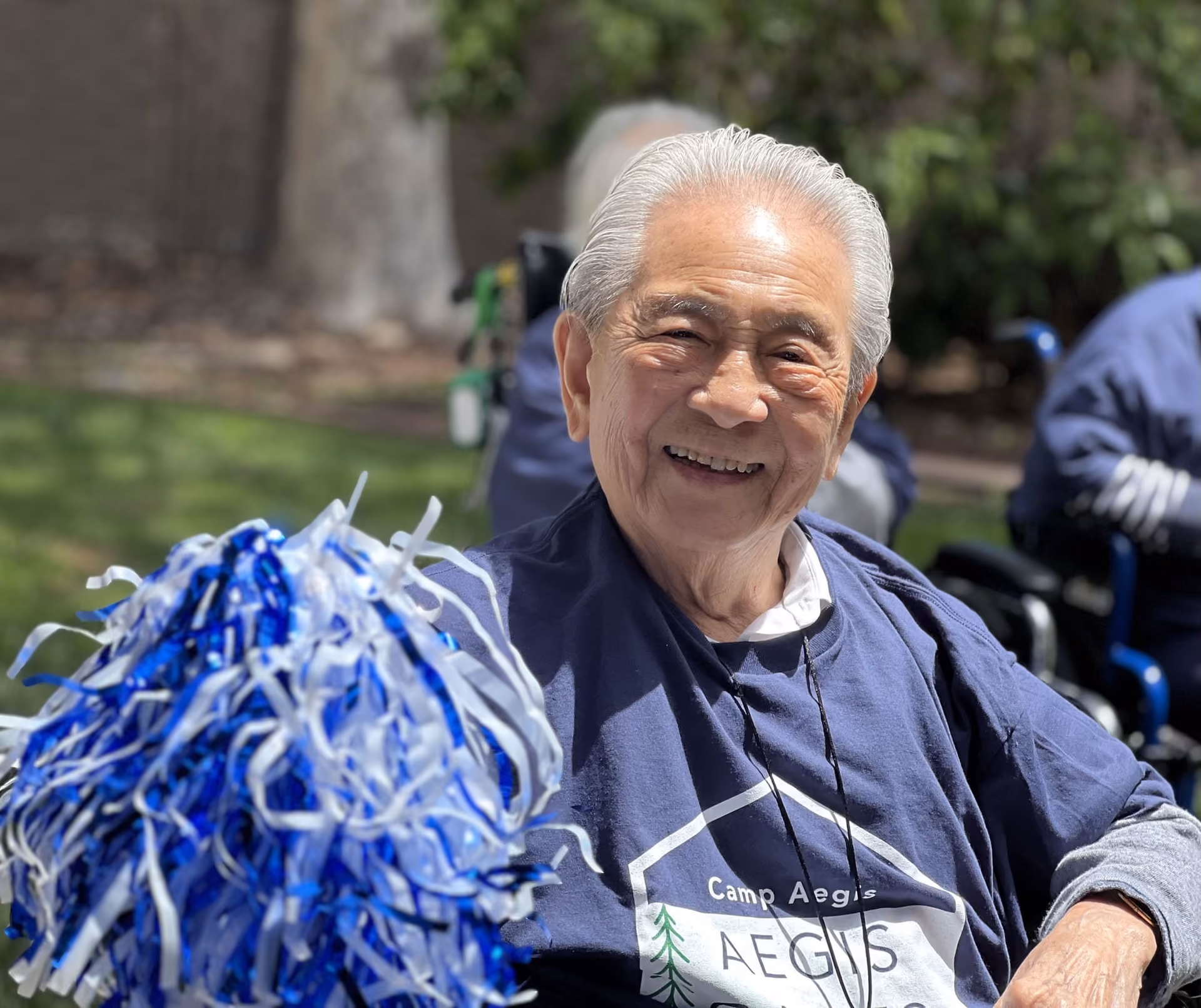 A smiling Áegis Gardens resident cheering with blue and white pom-poms during the Áegis Games
