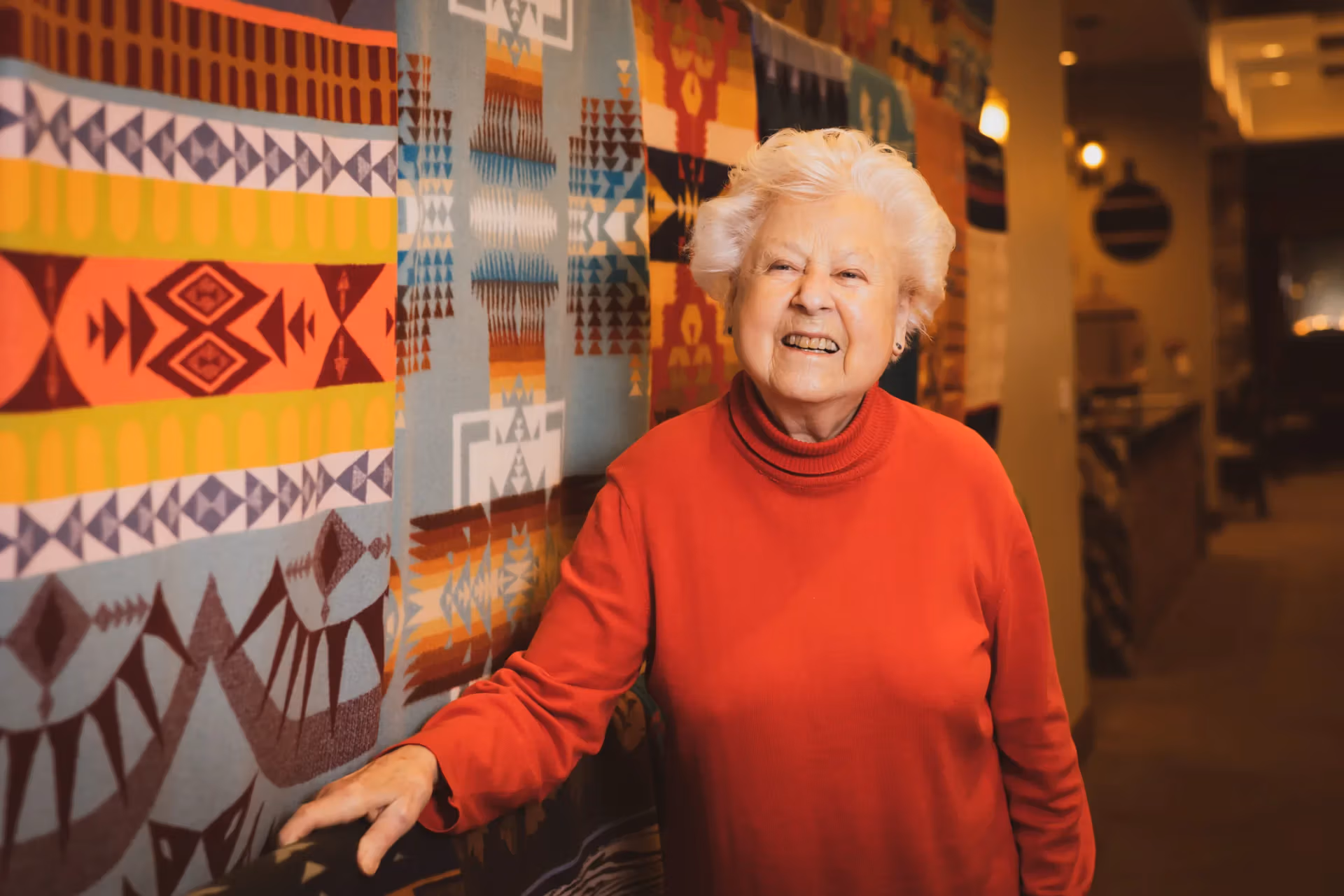 A resident smiling beside the warm textile art in a Life's Neighborhood common area
